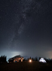 Colorful night starry sky and Milky Way over tourist camp set on mountain hill. Black SUV,...