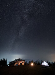 Colorful night starry sky and Milky Way over tourist camp set on mountain hill. Black SUV, illuminated tent and bonfire between them on background of silhouettes of trees and mountains. © anatoliy_gleb