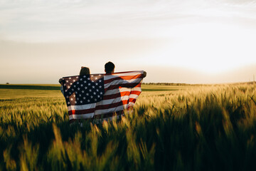 Patriotic holiday. Young couple with the American flag in a wheat field at sunset. Independence...