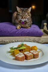 The cat sits on a purple cushion in front of a plate of smoked fish
