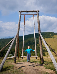 Blonde woman sitting on seat and putting hands on two support of giant street swing made from wooden frame, on background of mountain hill covered with trees.