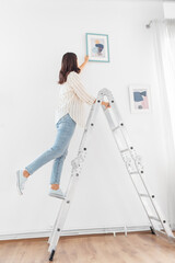 home improvement, decoration and people concept - happy smiling woman on ladder decorating home with art - obrazy, fototapety, plakaty