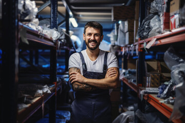 Young smiling happy tattooed bearded worker in overalls standing in storage of import and export...