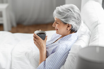 morning, old age and people concept - happy smiling senior woman in pajamas with cup of coffee sitting on bed at home bedroom