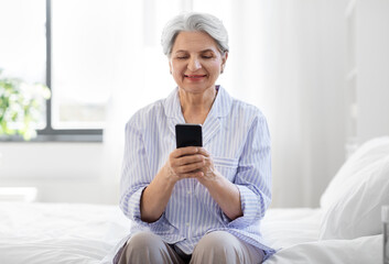 old age, technology and people concept - happy smiling senior woman in pajamas with smartphone sitting on bed at home bedroom