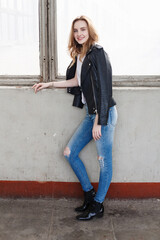 portrait of young caucasian woman standing in white t-shirt, black leather jacket and blue jeans at coridor with windows. pretty lady stands in room