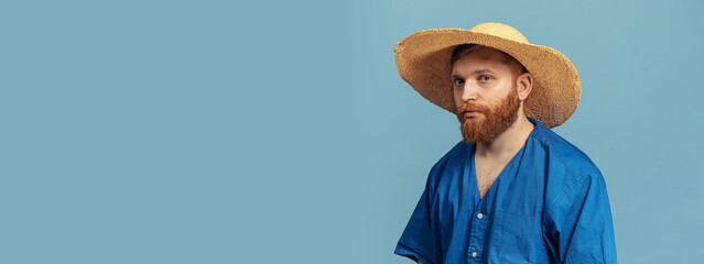 Young red headed and bearded man in straw hat standing isolated over blue bacground. Flyer
