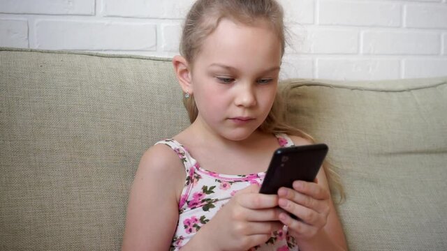 Girl playing mobile game on smartphone on sofa. Preschooler playing mobile phone. Kid using phone for gaming. Child playing video game at home