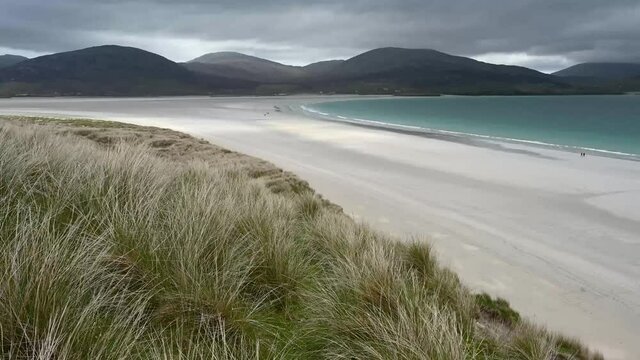 4k real time dune grass blowing in wind at Luskentyre beach, Isle of Harris. Moody sky with dark clouds, white sand beach, turquoise sea and mountains in background. Distant unidentifiable people.