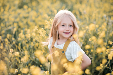 Naklejka na ściany i meble Funny blonde kid girl 4-5 year old wear yellow trendy dress posing in blooming meadow of rape flowers over nature background outdoors. Summer holiday season. Carefree childhood lifestyle. Happiness.