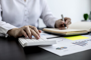 An accounting businesswoman pressing the white calculator and taking notes in her notebook, she was examining the company's financial accounts from the documents provided by the finance department.
