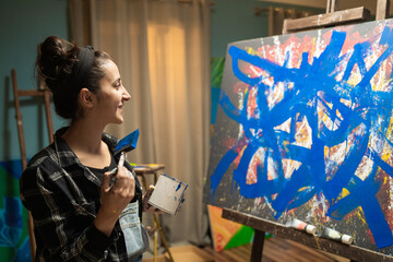 A talented beautiful artist is working on an abstract oil painting, looking at her modern masterpiece with a broad smile. In the studio stand easels, canvases, daylight.