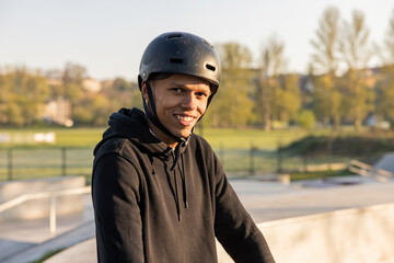 A teenager in a black sweatshirt and helmet stands on a ramp at a skatepark and smiles for the...