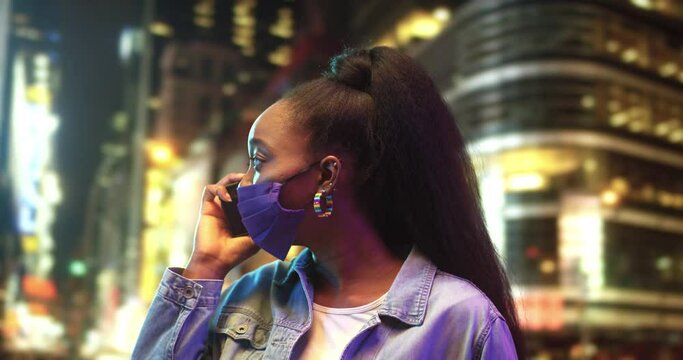 Be on touch. Portrait view of the gorgeous multiracial woman wearing protective mask holding smartphone and chatting with somebody while making spending time at the night city