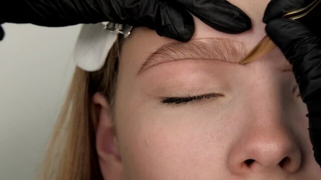 Gloved master's hands pluck model's eyebrows with tweezers close-up. Eyebrow lamination and styling. Shape of the eyebrows is drawn with a pencil on the face of a beautiful young woman. 