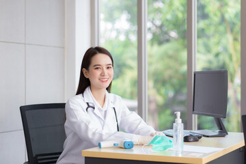 Asian beautiful young smiling female doctor wears rubber glove sitting in office at hospital. On table has a paper,face mask ,alcohol gel ,temperature meter and computer.