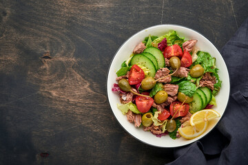 Tuna salad with fresh vegetables, olives, capers and lemon served in bowl on old wooden background. Top view with copy space.