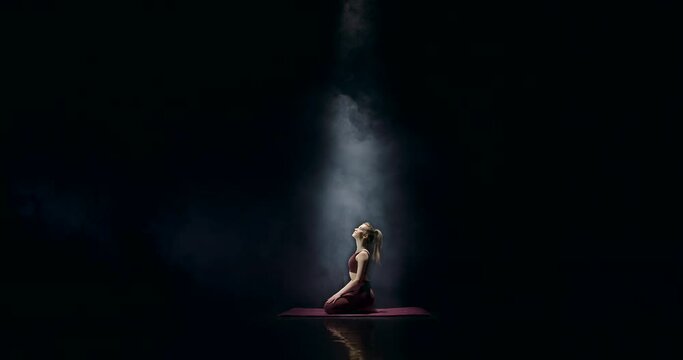 In a dark auditorium, a young and attractive woman sits on a gymnastic rug on stage. She meditates and stretches the muscles in her legs. She slowly lifts her head up and looks at the beam of light