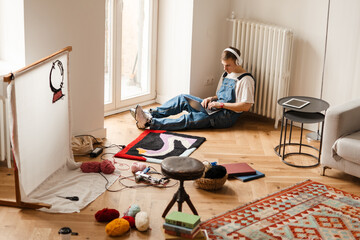 Young man in headphones using computer while working on craft rug
