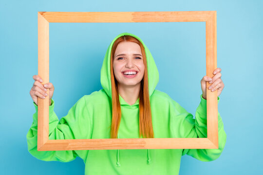Photo of pretty sweet young woman wear green sweatshirt hood holding wooden frame smiling isolated blue color background