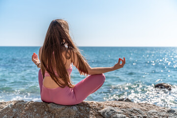 A young woman in gray leggings and a pink top with long loose hair does yoga outdoors by the sea on a sunny day. Women's yoga, fitness, Pilates. The concept of a healthy lifestyle, harmony.
