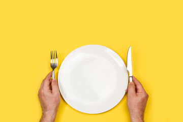 Manos de hombre sosteniendo unos cubiertos junto a un plato redondo blanco vacío sobre un fondo amarillo liso y aislado. Vista superior. Copy space