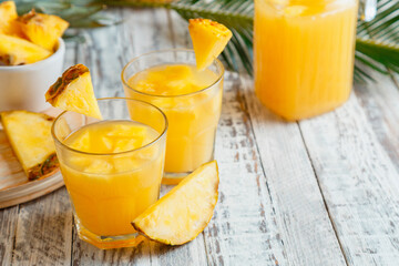 Tasty pineapple juice in two glasses with pineapple slices. Fresh natural pineapple cocktail and juice in Glass Jug on white wooden table with palm leaves.