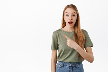 Fototapeta premium Wow super. Excited and curious girl pointing left with opened mouth, showing awesome new promo deal, store banner on copyspace, standing over white background