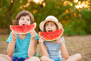 Two preschool smiling child sister sit with crossed legs on grass and eating watermelon at summer park with sunset light