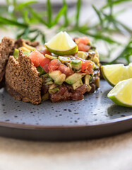 Raw tuna and avocado tartar served with rye bread crackers and lime on black ceramic plate. Gourmet fish tartar.