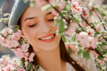 Fototapeta premium Happy smiling beautiful girl with natural makeup posing near blooming sakura flowers. Model closed eyes. Close up outdoor portrait. Spring, beauty, skin care, conception. 