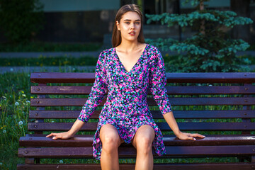 Young brunette woman in a lilac summer dress posing while sitting on a park bench