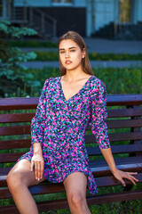 Fototapeta premium Young brunette woman in a lilac summer dress posing while sitting on a park bench