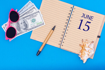 Travel concept flat lay - notepad with the date of 15 june pen, glasses, dollars and seashell on blue background.
