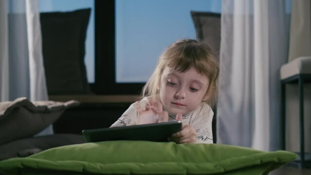 Curious Cute Preschool Kid Girl Using Digital Tablet Technology Device Lying On Sofa Alone. Small Child Hold Pad Computer Surfing Internet Play Game At Home. Children Tech Addiction Concept