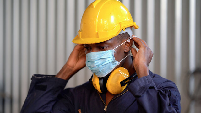 African American Worker Man Wearing Protective Face Mask In Industry Factory During Coronavirus Or Covid 19 Pandemic   . Black Engineer Putting Medical Mask On Face Against Air Pollution Indoors .