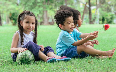 Three little mixed race kids consist of African and caucasian boys and girls smiling with happiness, fun amusement, playing, sitting for picnic and eating piece of watermelon fruit in outdoor garden.