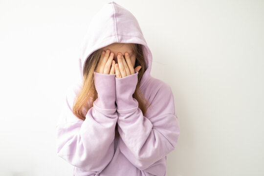 close up hooded upset girl covers her face with her hands standing against white wall