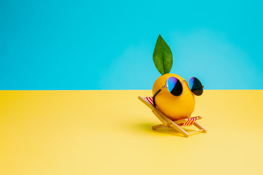 Lemon fruit chilling in beach chair on the blue and yellow background. Summer vacation concept. Sunglasses on lemon with green leaf relaxing on the sunbed. Creative art, minimal aesthetic.