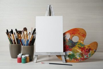 Easel with blank canvas, brushes, paints and palette on white wooden table
