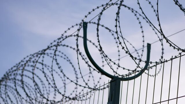 Barbed wire Hanging on the Border of Iron Fence on Against the Blue Sky. Restricted area. Barrier from strangers people, refugees, prisoners. Lattice. Jail. 4K. Close up.