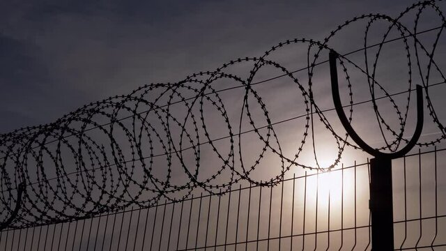 Barbed Wire Hangs on Border of an Iron Fence Against the Backdrop of Sunset. Restricted area. Barrier from strangers people, refugees, prisoners. Lattice. Jail. 4K. Close up.