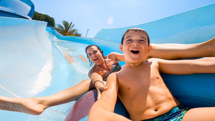 Happy cheerful caucasian son and mom laughing and shouting while sliding down fast on a water slide...