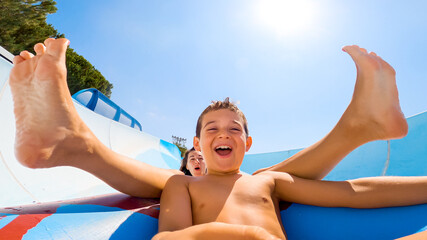 Smiling happy boy and mom sliding down fast on a rubber inflatable ring through a water slide at...