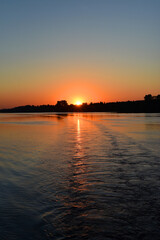 Naklejka premium Early morning. Sunrise. View from the river with the reflection of the shore in the water and the wake of the ship. Vertical