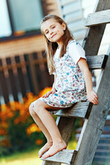 A beautiful country girl with loose hair in a summer sundress sits on a wooden staircase and looks at the camera. A young schoolgirl is resting on a summer day in the village.