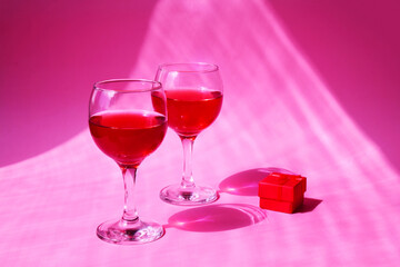 Two glasses with a red gift box on a pink background with sun glare and hard light. Beautiful background, concept for February 14, symbol of love or romantic date