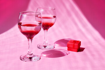 Two glasses with a red gift box on a pink background with sun glare and hard light. Beautiful background, concept for February 14, symbol of love or romantic date