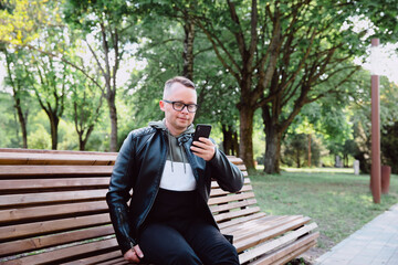 A young Caucasian man in black jeans and a leather jacket sits on a bench in a city park, reads the news via the Internet on his smart phone, communicates via video link.