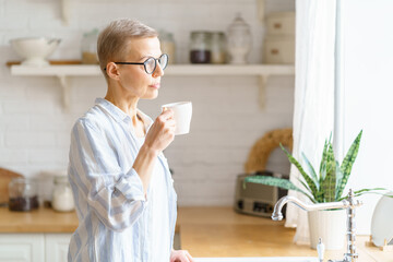 Enjoying new day. Thoughtful beautiful mature woman holding cup of coffee or tea, pure water,...